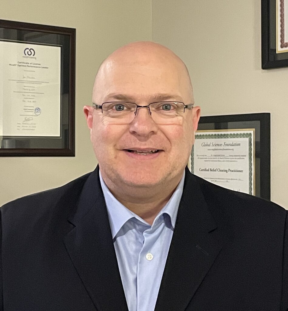 Headshot photo of Joe Trevors, serial entrepreneur, author, and Functional Wellness Practitioner dressed in a light-blue dress-shirt, navy suit coat and glasses, standing in front of a wall with a few certificates hanging in frames.