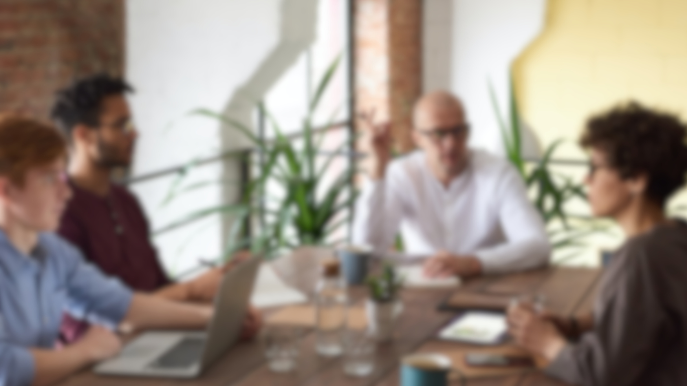 A somewhat blurred picture of what appears to be an exclusive agency meeting room, with 4 people situated around the table. The person speaking appears to be a bald man with glasses, wearing a white shirt.