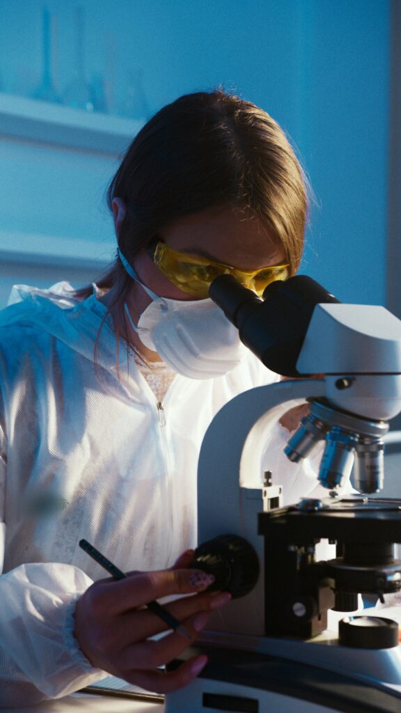 A female genetic scientist - dressed in a white lab suit, mask, and protective glasses views through the eye-lens of a laboratory microscope.