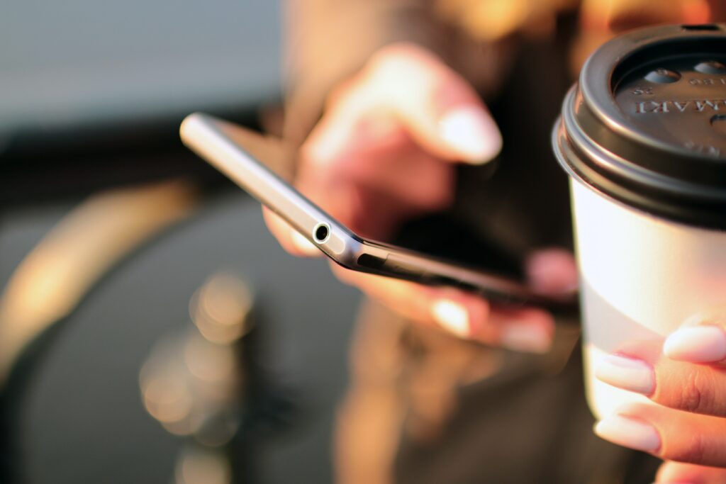 A zoomed-in picture of someone's hands. iPhone in one hand, white paper coffee cut with a brown plastic lid on the other.