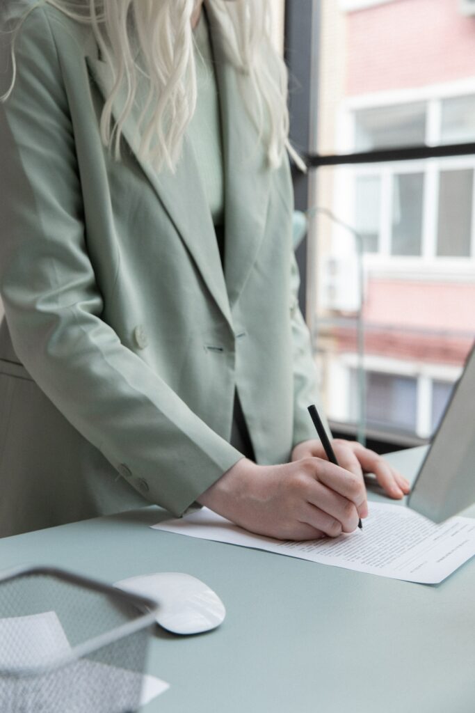 Picture of a tall, lean, sharply-dressed ad agency woman in a light-grey coat. Pen in hand, writing on a piece of paper beside an open MacBook at a standing desk.
