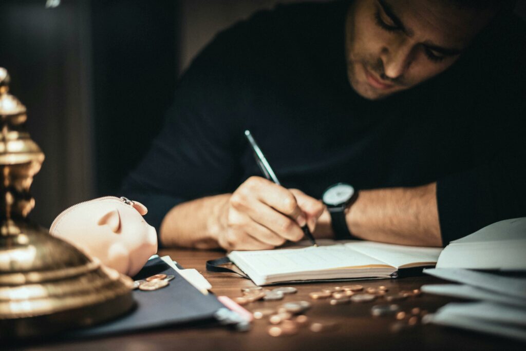 A man in a black sweater sits at a desk, writing in a journal or ledger, with a gold lamp, a pink piggy bank, and scattered coins on the desk.