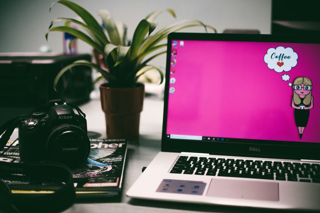 Desk setup with Dell laptop, photography camera, magazines, and plant, with cartoon character in thought bubble asking "Coffee?"