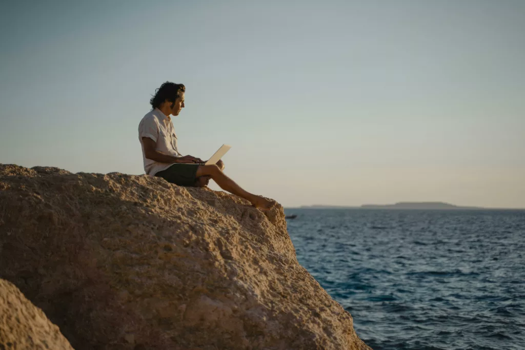 A man in a tropical shirt and cargo shorts works on a laptop while overlooking the ocean from a rocky outcrop.